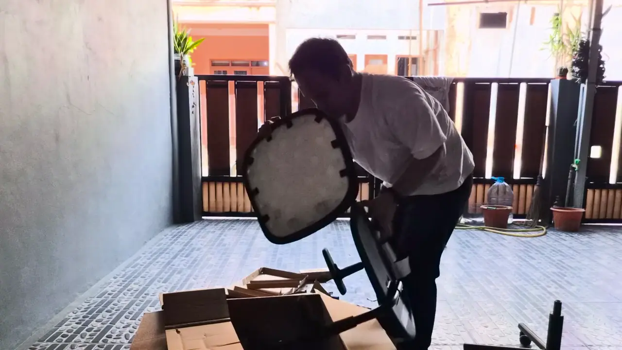 Holding the seat shell during assembly, with the underside of the seat visible and the flat-pack box on the ground